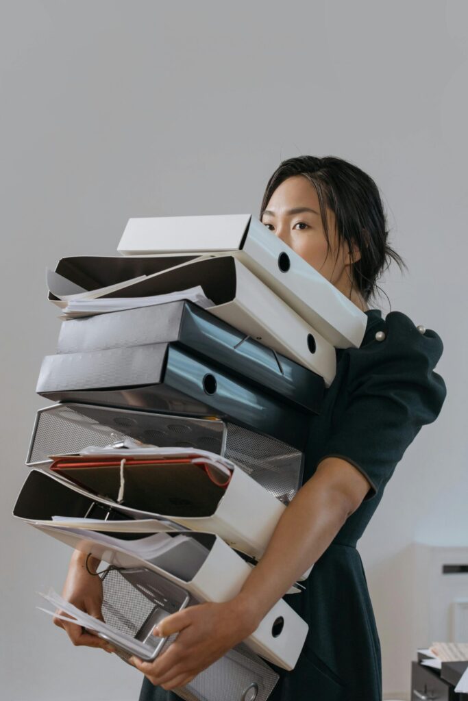 Woman carrying a large stack of file folders indoors, expressing stress and workload.